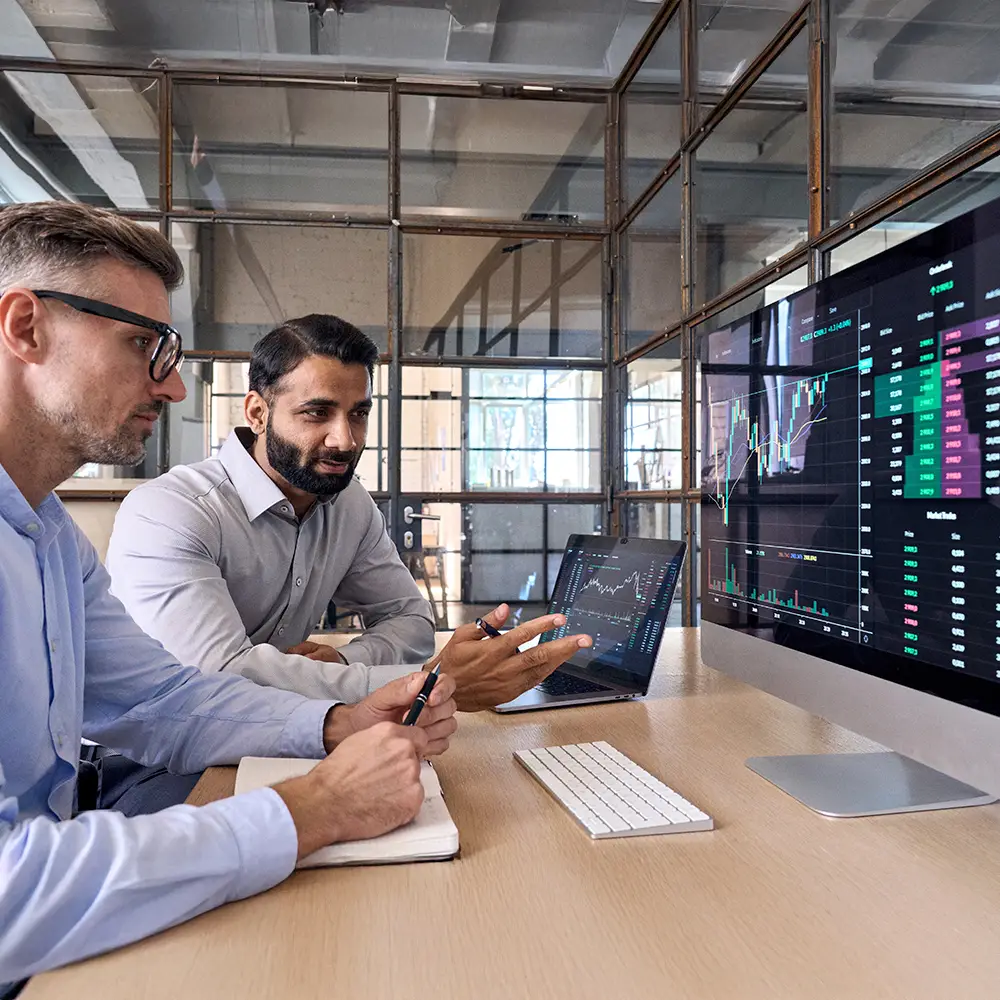 Two professionals analysing cryptocurrency market data on large monitors and a laptop in a modern office.