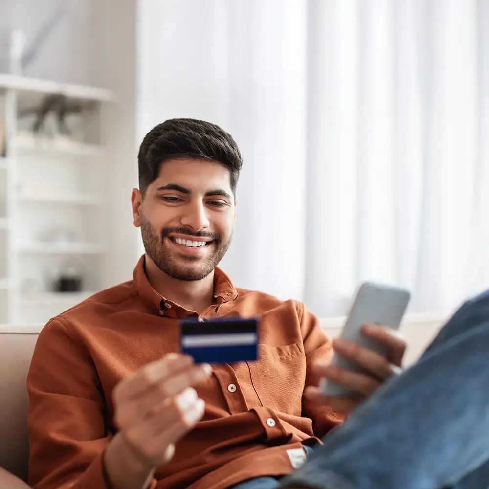 Man sitting on a sofa smiling while making an online purchase with a credit card and smartphone.