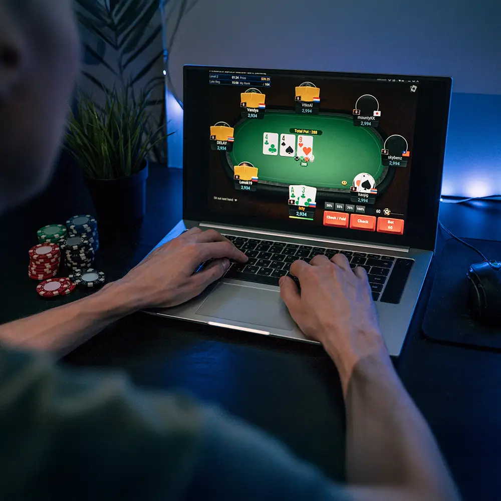 Person playing online poker on a laptop, surrounded by gaming chips in a dimly lit setup.