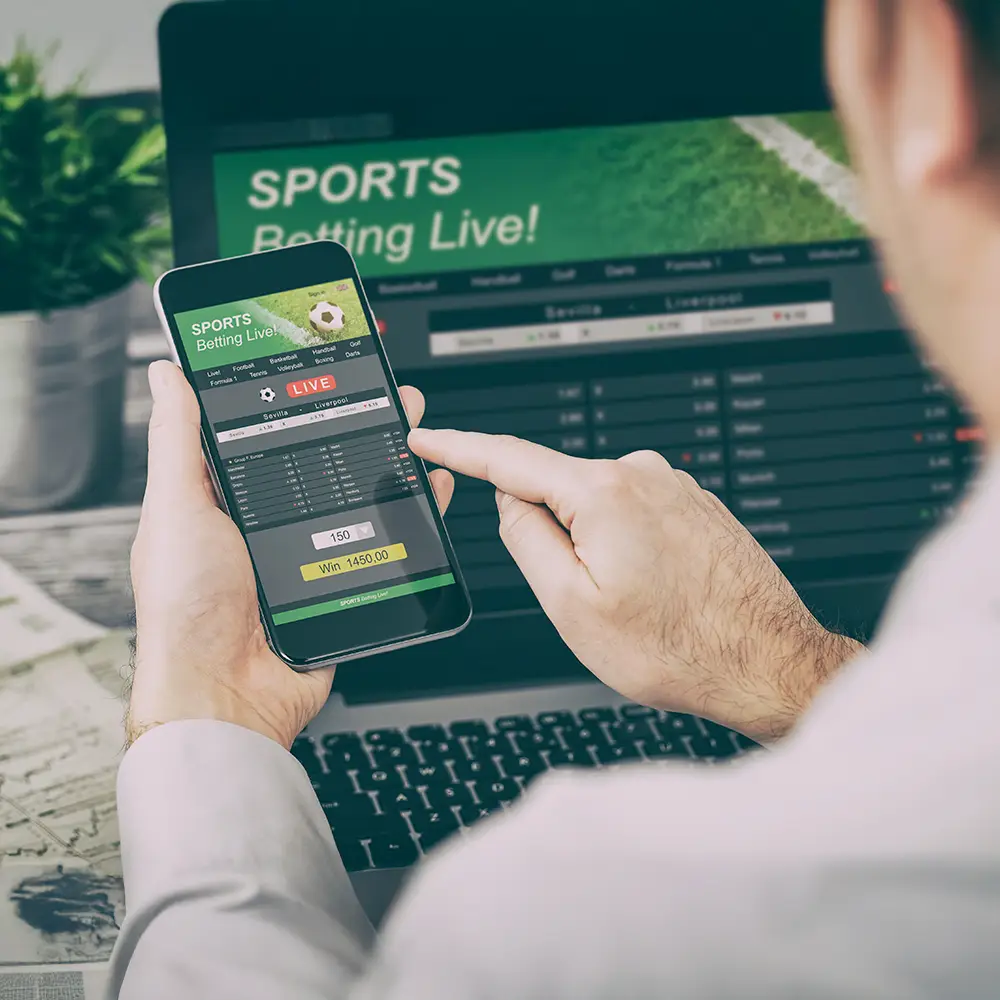 User placing a live sports bet through a mobile app while reviewing betting statistics on a laptop screen.