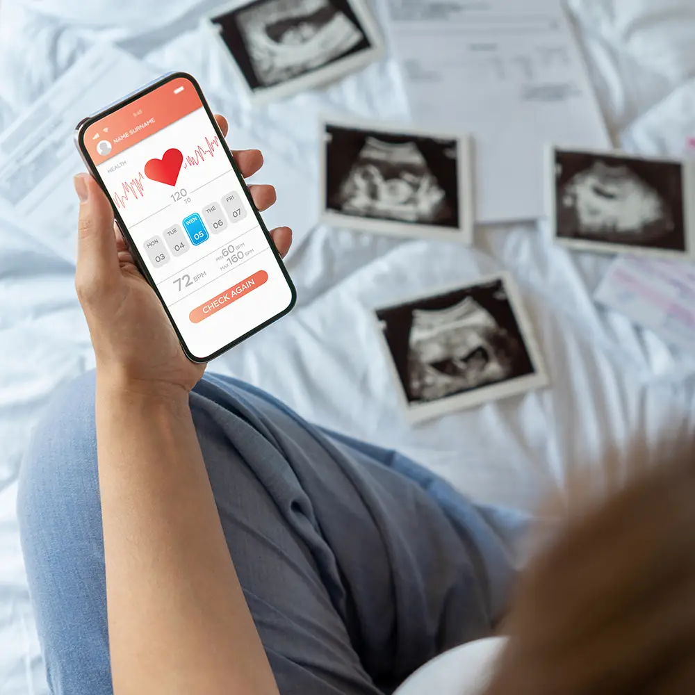 Pregnant woman holding a smartphone with a healthcare app, surrounded by ultrasound images on a bed