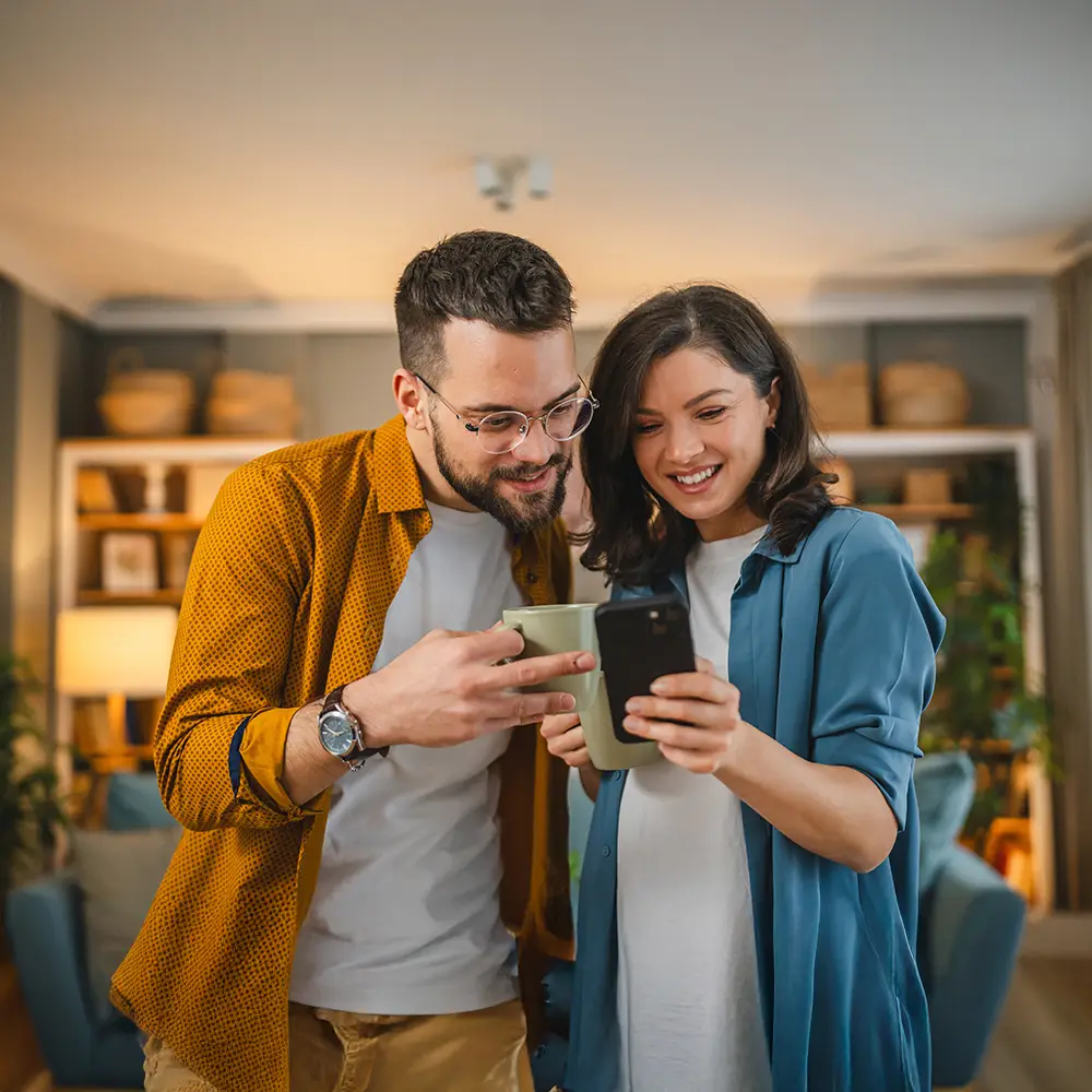 Couple at home smiling while looking at a smartphone together, representing modern telecommunications and digital connectivity.